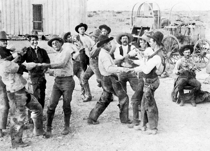 Photograph of cowboys dancing to a fiddle (far right) at the Bar S Ranch, which was located in Reagan and Irion counties out in west Texas. A wagon and a building stand in the background.